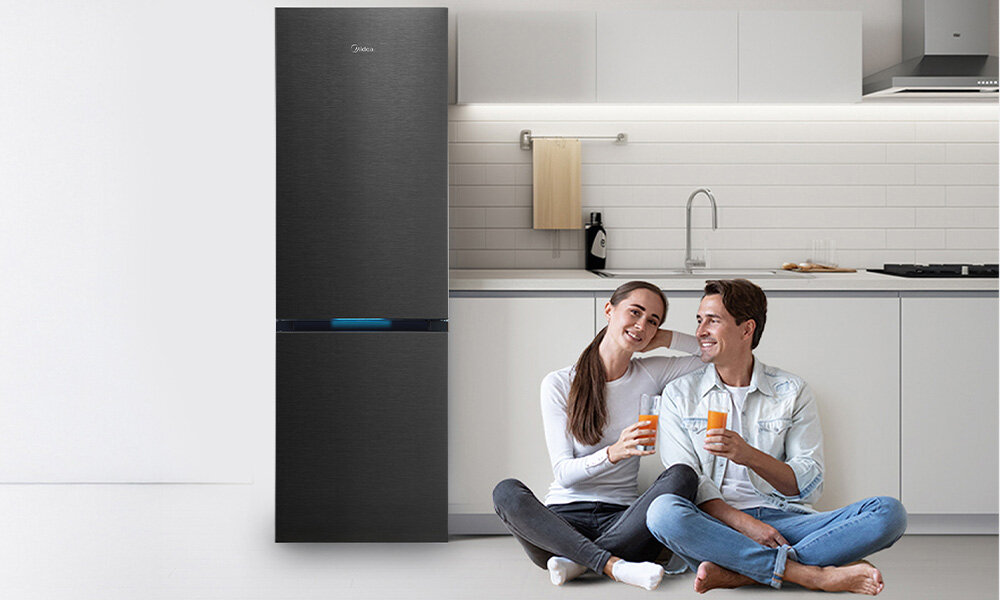 LODÓWKA MIDEA MDRB548MIC45P Para siedzi na podłodze w kuchni, trzymając napoje i spoglądając na siebie z uśmiechem. Obok nich stoi lodówka w ciemnym kolorze, wkomponowana w jasne wnętrze minimalistyczny design elegancki kolor wnętrze AGD
