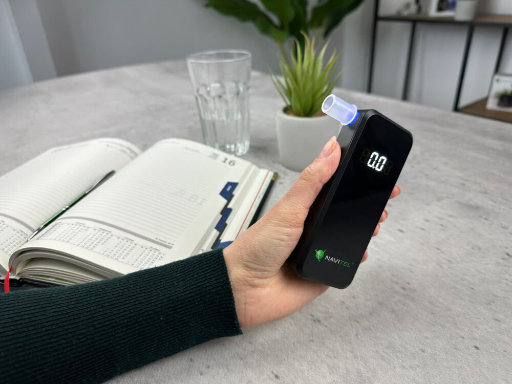 NAVITEL ALC05 NAVITEL ALC05 Pro zawartość alkoholu w wydychanym powietrzu elektrochemiczny sensor 0,00 do 4,00‰ 0,01‰ - Pomiar poziomu alkoholu przy użyciu alkomatu, kobieta trzyma urządzenie na tle biurka z kalendarzem