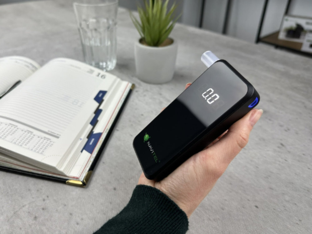NAVITEL ALC05 NAVITEL ALC05 Pro 118 x 54 x 20 mm 73 g kieszeni kurtki, damskiej torebce czy wewnętrznej kieszeni marynarki etui - Widok alkomatu podczas testu, na biurku obok szklanki i rośliny doniczkowej