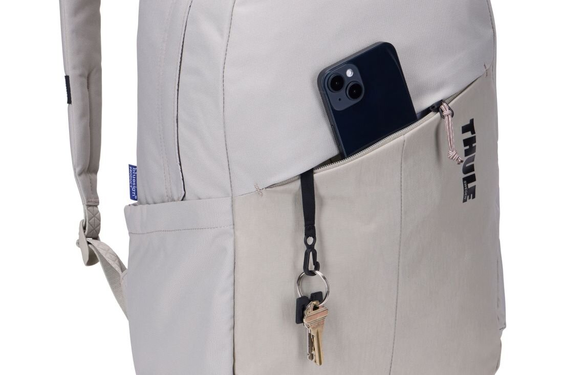 Jasnobeżowy plecak z widoczną kieszenią, w której znajduje się telefon i przypięte klucze. Na kieszeni widoczne jest logo \'THULE\'.