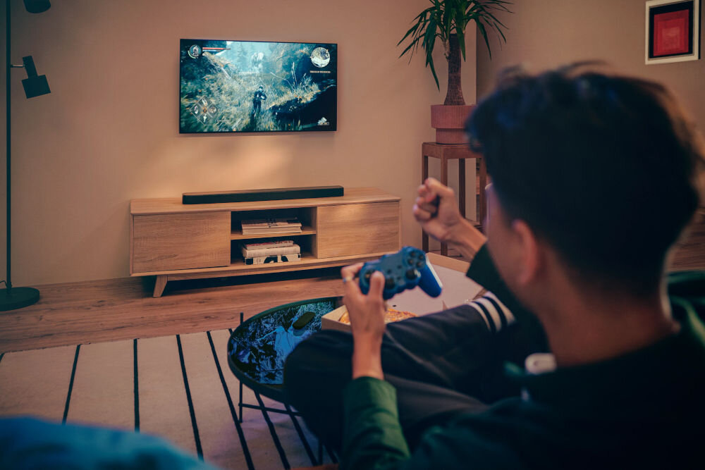 Soundbar JBL BAR 300 MkII kalibracja akustyki automatyczne dostosowanie efektów przestrzennych 3D, osoba trzymająca kontroler w ręku, grająca w grę wideo na zawieszonym telewizorze