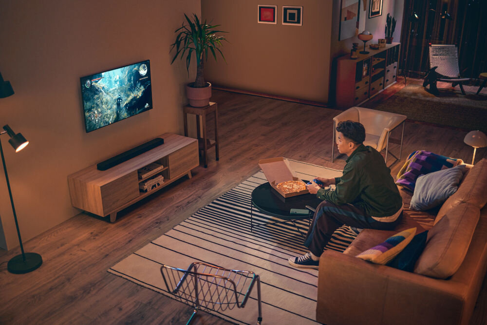 Soundbar JBL BAR 300 MkII technologia Dolby Atmos dźwięk 3D kino domowe, osoba trzymająca kontroler w rękach, grająca w grę wideo na zawieszonym telewizorze