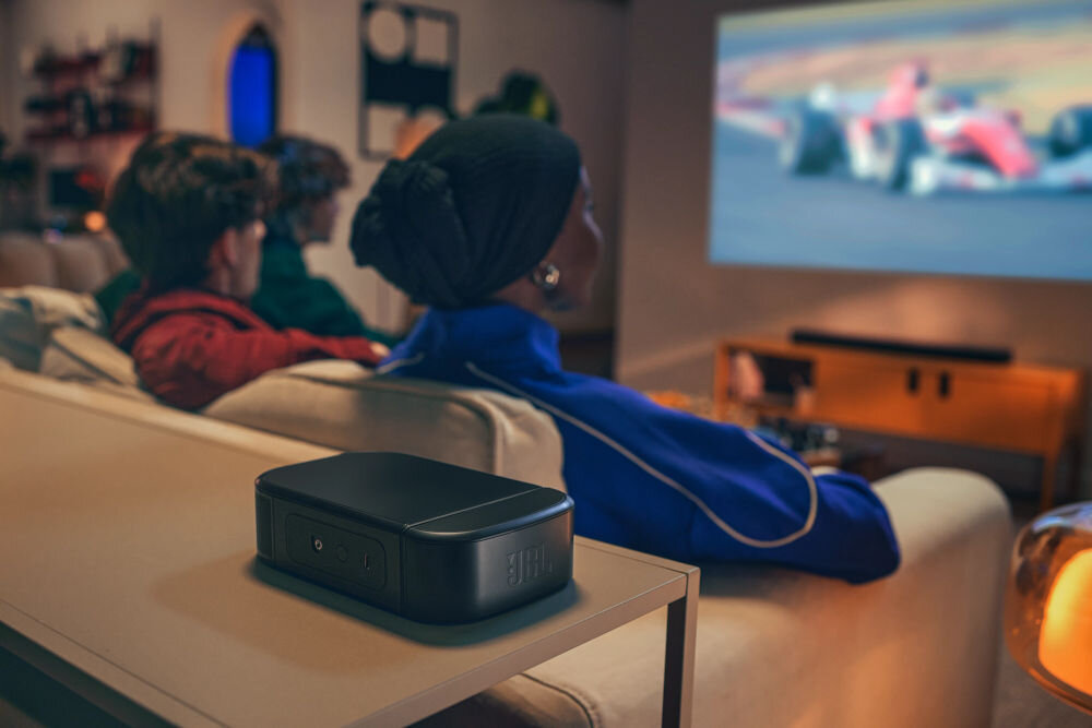 Soundbar JBL BAR 800 MkII PureVoice 2.0 wyraźne dialogi optymalizacja, głosnik surround sotjący na stoliku, w tle osoby oglądające formułe 1