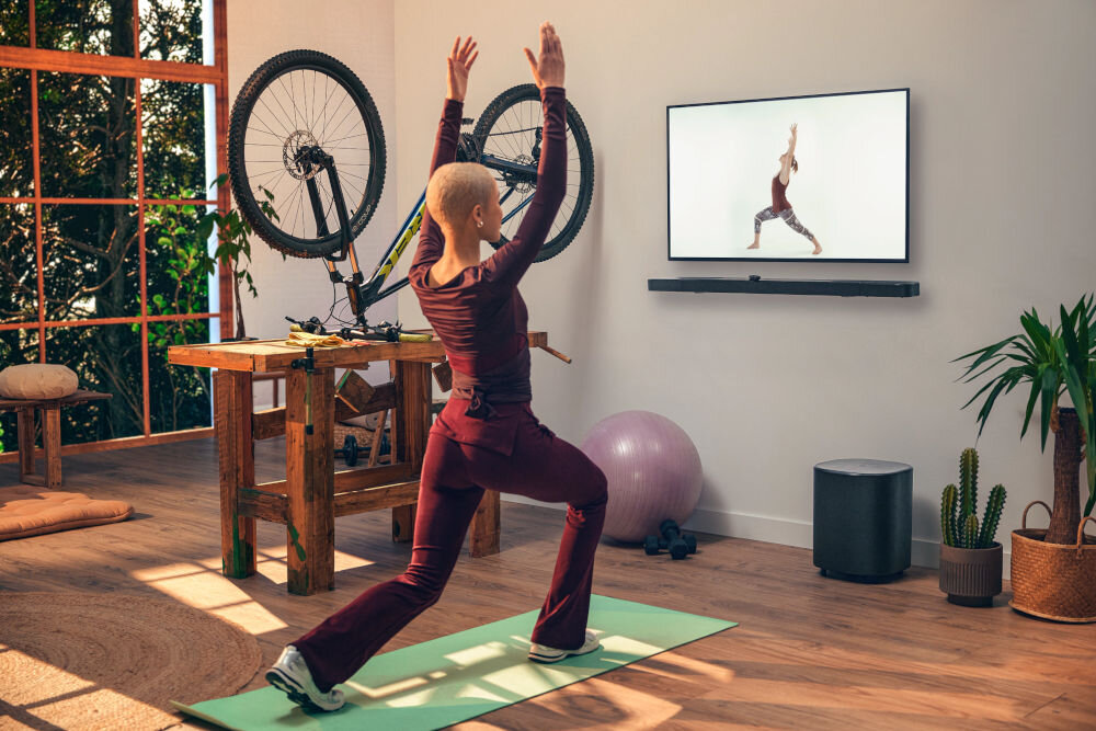 Soundbar JBL BAR 1000 MkII obsługa głosowa Google Assistant AI Detail Enhancer Sound Boost, osoba ćwicząca przed włączonym telewizorem