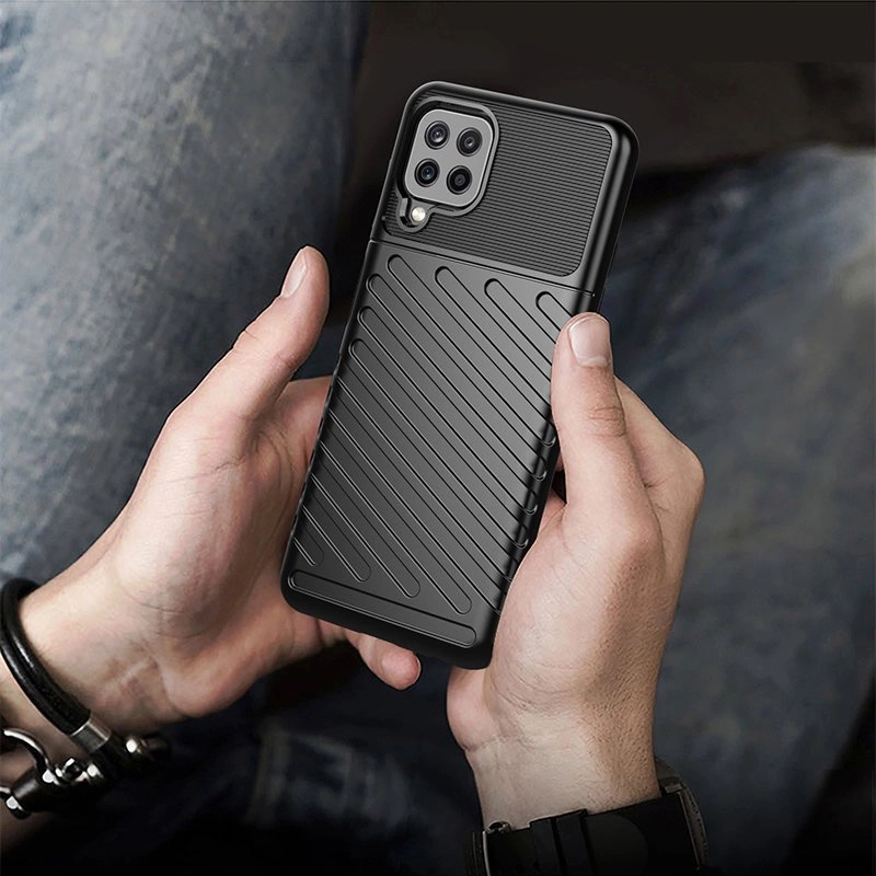 Osoba trzyma telefon w czarnym, pancerzowym etui z wyraźnymi, ukośnymi żłobieniami. Etui ma wycięcia na aparat z czterema obiektywami.