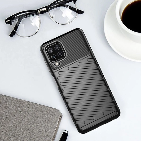 Na obrazie widoczne jest czarne etui na telefon z teksturowanym wzorem, leżące na szarej powierzchni obok okularów, filiżanki z napojem i notesu. Etui ma wycięcia na aparat i przyciski telefonu.