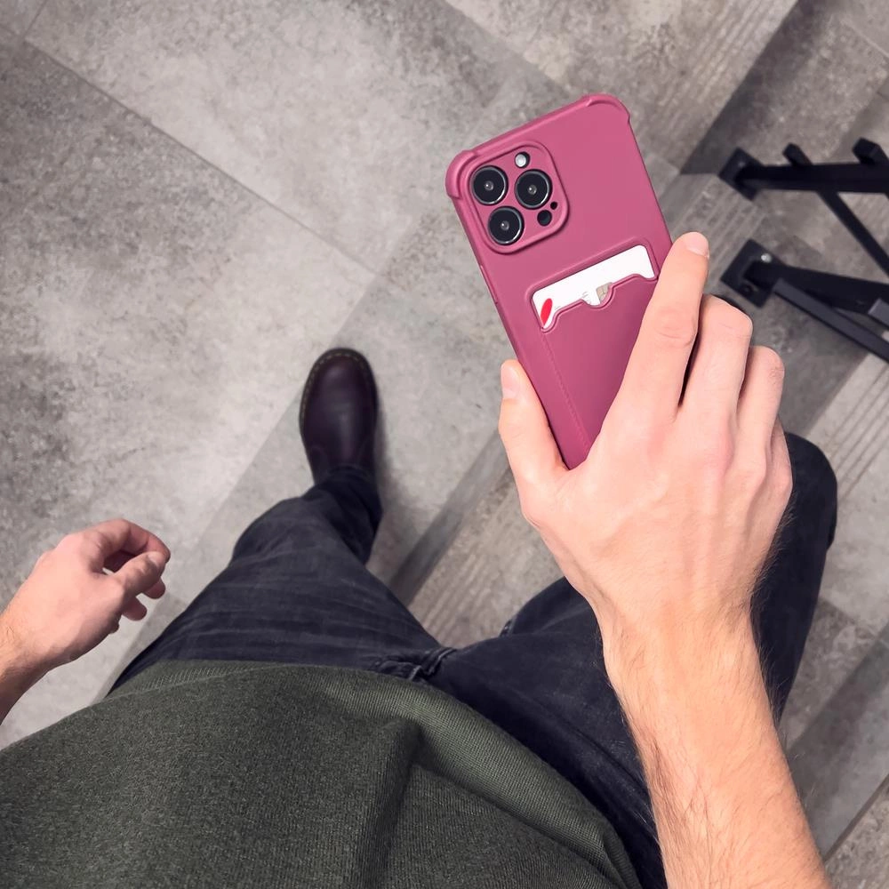 Osoba trzyma malinowe etui na telefon z miejscem na kartę. W tle widać szare płytki podłogowe.