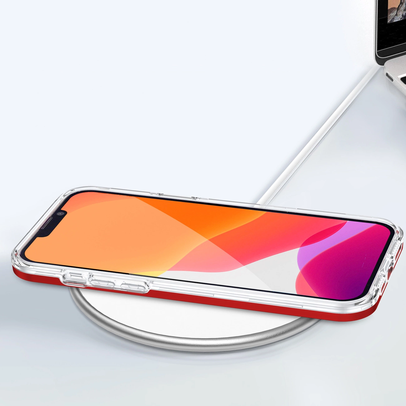 Na obrazie widać telefon w przezroczystym etui z czerwoną ramką, leżący na białej, okrągłej ładowarce bezprzewodowej. W tle znajduje się fragment otwartego laptopa.