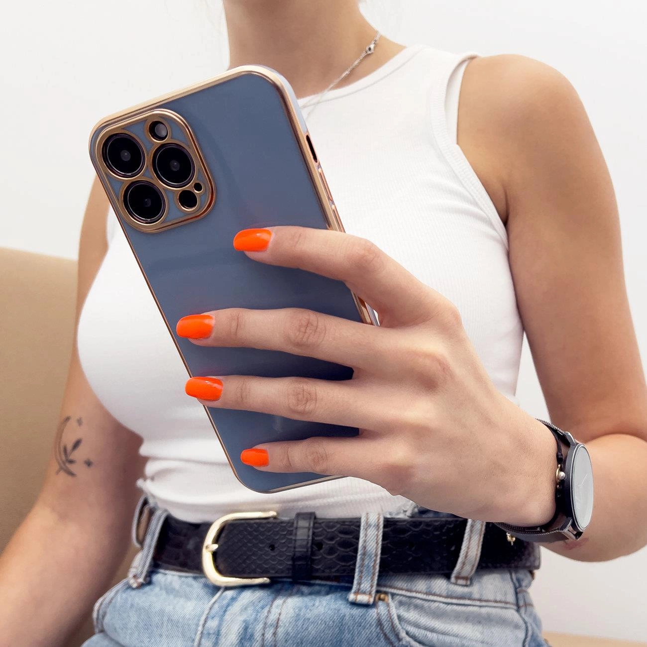 Osoba trzyma telefon w etui z niebieskim tyłem i złotą ramką, z widocznymi trzema aparatami. Osoba ma pomalowane na pomarańczowo paznokcie i nosi biały top oraz zegarek na czarnym pasku.