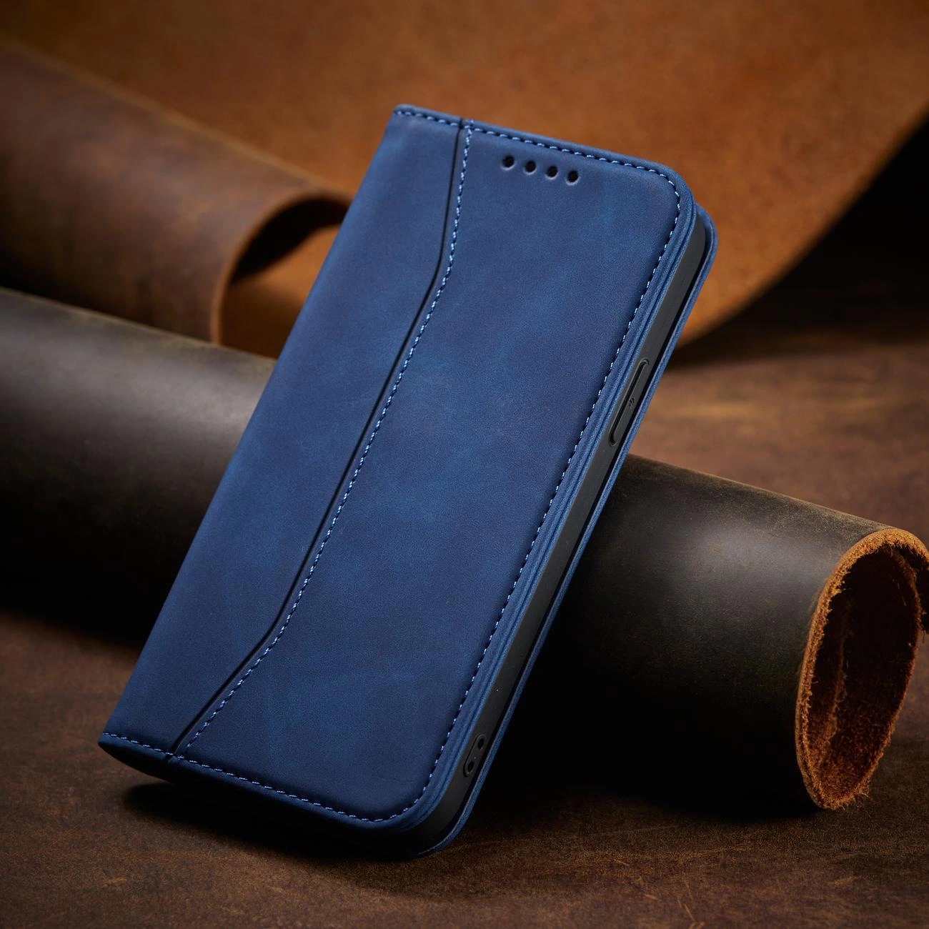 Niebieskie etui na telefon z klapką, wykonane z materiału przypominającego skórę, z widocznymi przeszyciami i otworami na głośnik. Etui jest ustawione na tle ciemnych, skórzanych materiałów.