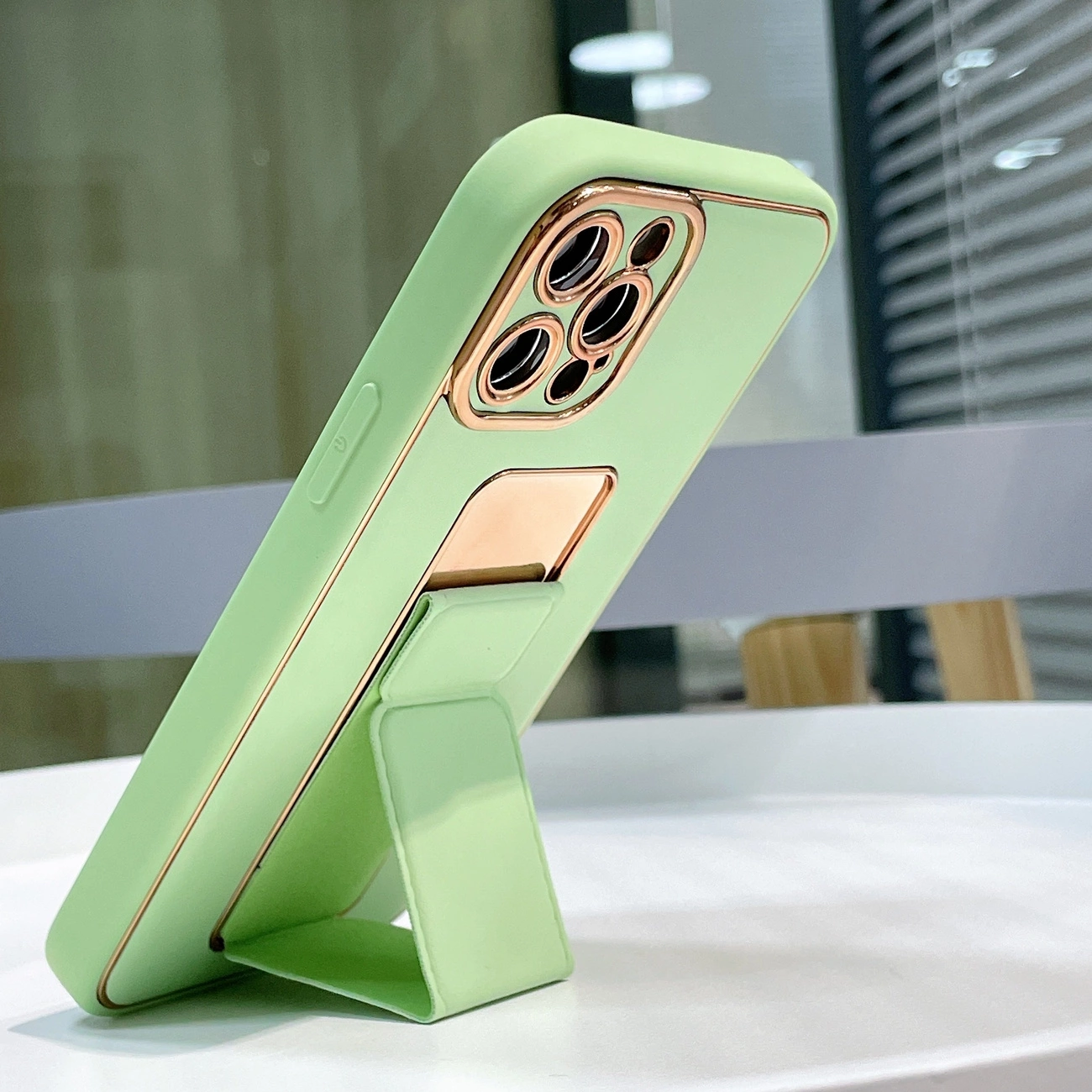 Zielone etui na telefon z podstawką, posiadające złote wykończenia wokół aparatu i podstawki. Telefon stoi na białej powierzchni, a w tle widać rozmyte elementy wnętrza.