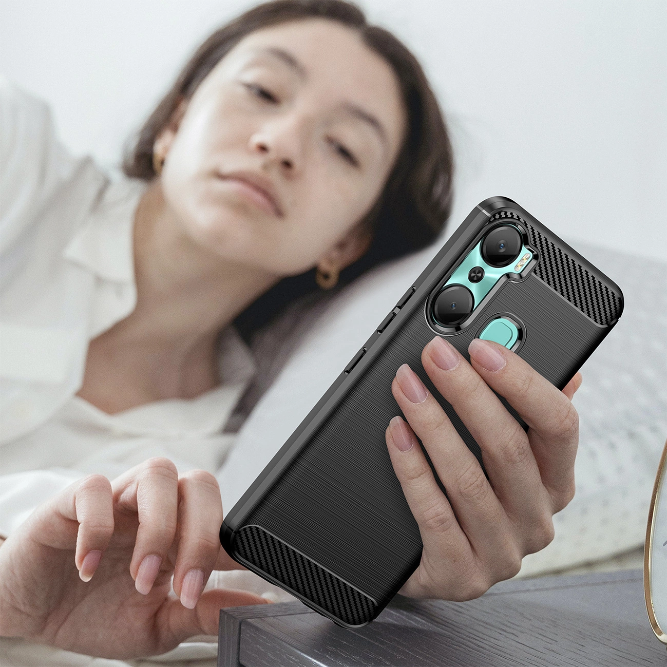 Osoba trzyma telefon w czarnym, karbonowym etui z widocznymi przyciskami bocznymi i wycięciem na aparat. W tle widać osobę leżącą na łóżku.