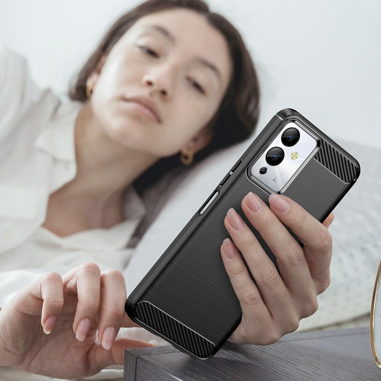 Osoba trzyma telefon w czarnym, karbonowym etui z widocznymi aparatami i przyciskiem bocznym. W tle widać rozmytą postać w jasnym ubraniu.