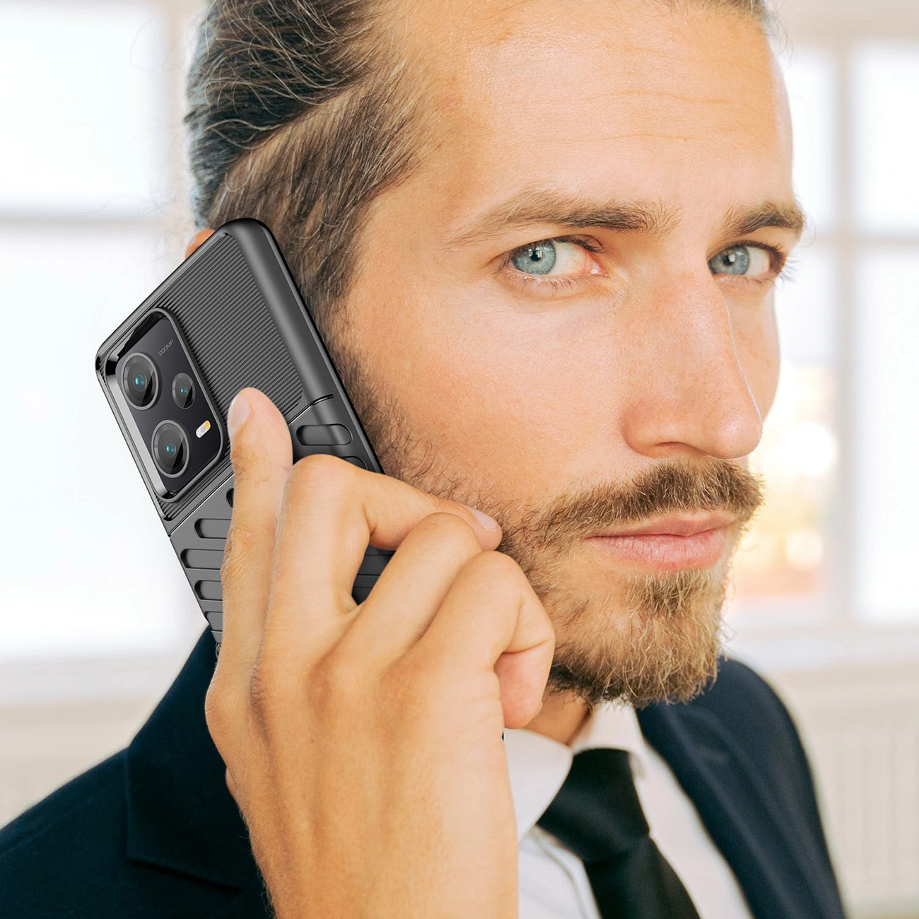 Mężczyzna trzyma przy uchu telefon w czarnym, pancernym etui z widocznymi trzema obiektywami aparatu. W tle znajduje się jasne wnętrze z dużym oknem.