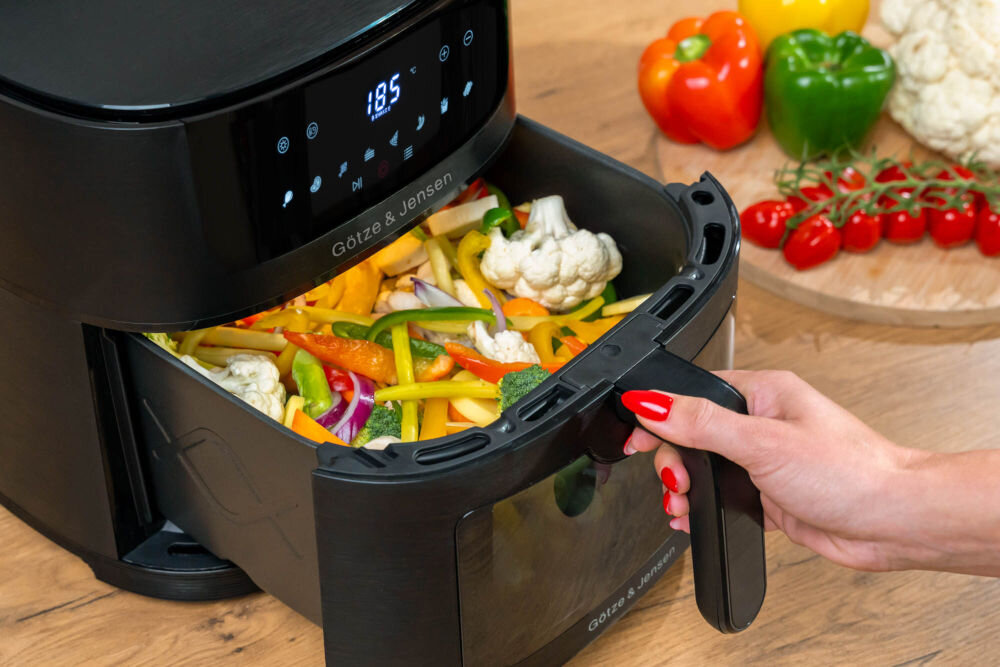 Frytkownica beztłuszczowa GÖTZE & JENSEN AF610K z nienagrzewającym uchwytem antypoślizgowymi nóżkami i zabezpieczeniem przed przegrzaniem Dłoń wysuwająca szufladę frytkownicy Götze & Jensen wypełnioną mieszanką warzyw, podkreślająca dużą pojemność 6 litrów.