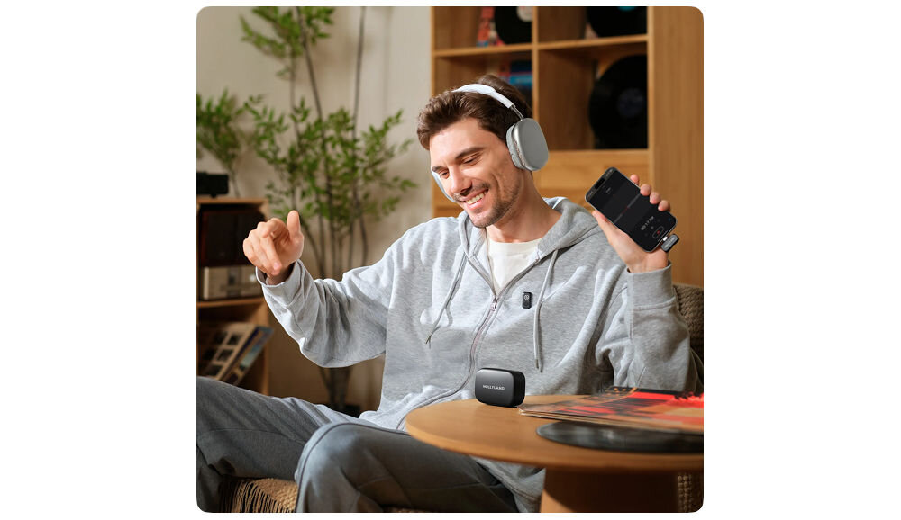System bezprzewodowy HOLLYLAND Lark A1 Mini Duo USB-C Szary prezentacja zestawu podczas użytkowania uśmiechnięty mężczyzna siedzący w pokoju w słuchawkach trzymający telefon etui na stoliku stosunek sygnału do szumu wysoka jakość nagrywanego dźwięku tłumi niepożądane zakłócenia