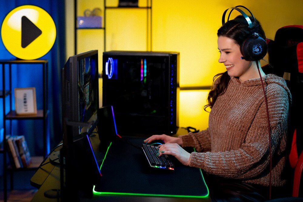 Komputer MAD DOG DEEPCOOL-A07WR64WP Kobieta w słuchawkach grająca przy biurku z dwoma monitorami i podświetlaną klawiaturą, w tle obudowa komputera z podświetleniem RGB, cechy, podzespoły, testy