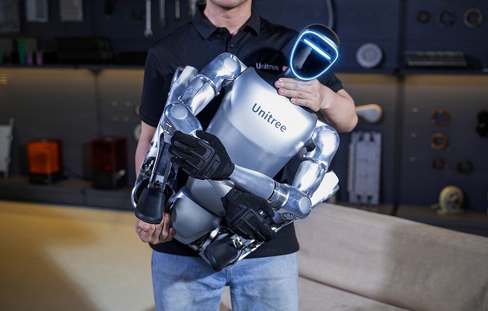 Osoba trzyma humanoidalnego robota z napisem \'Unitree\' na klatce piersiowej. Robot ma srebrne elementy i świecącą linię na twarzy.