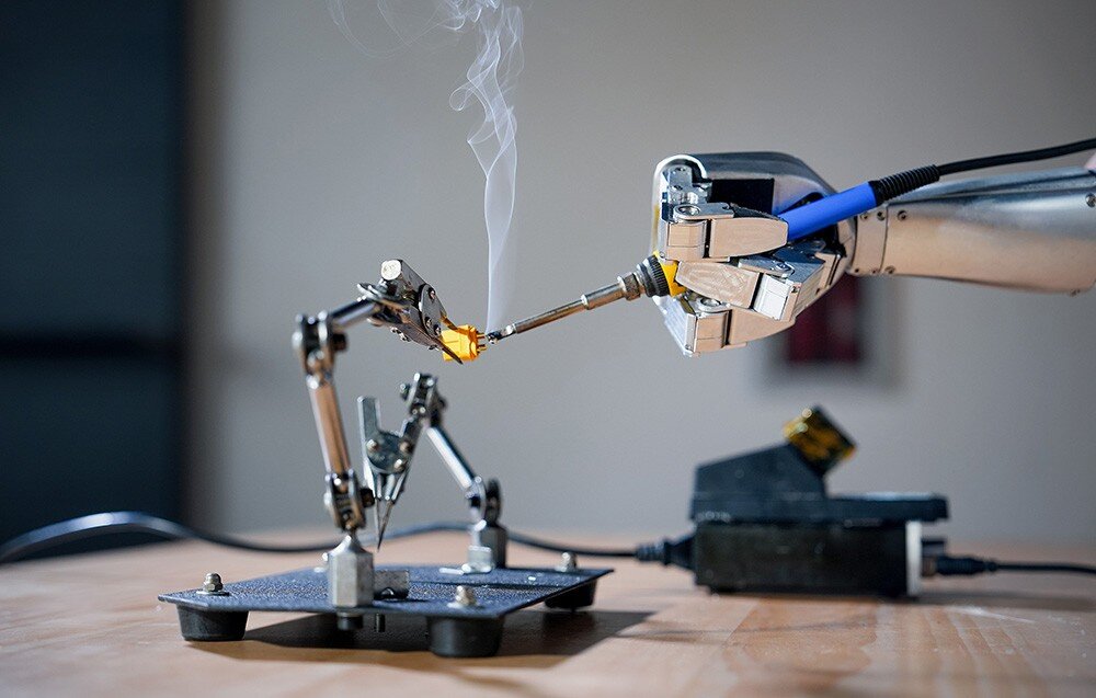 Na zdjęciu widać ramię robota trzymające lutownicę, która dotyka elementu elektronicznego zamocowanego na uchwycie. W tle znajduje się rozmyty element, a na pierwszym planie widoczna jest powierzchnia stołu.