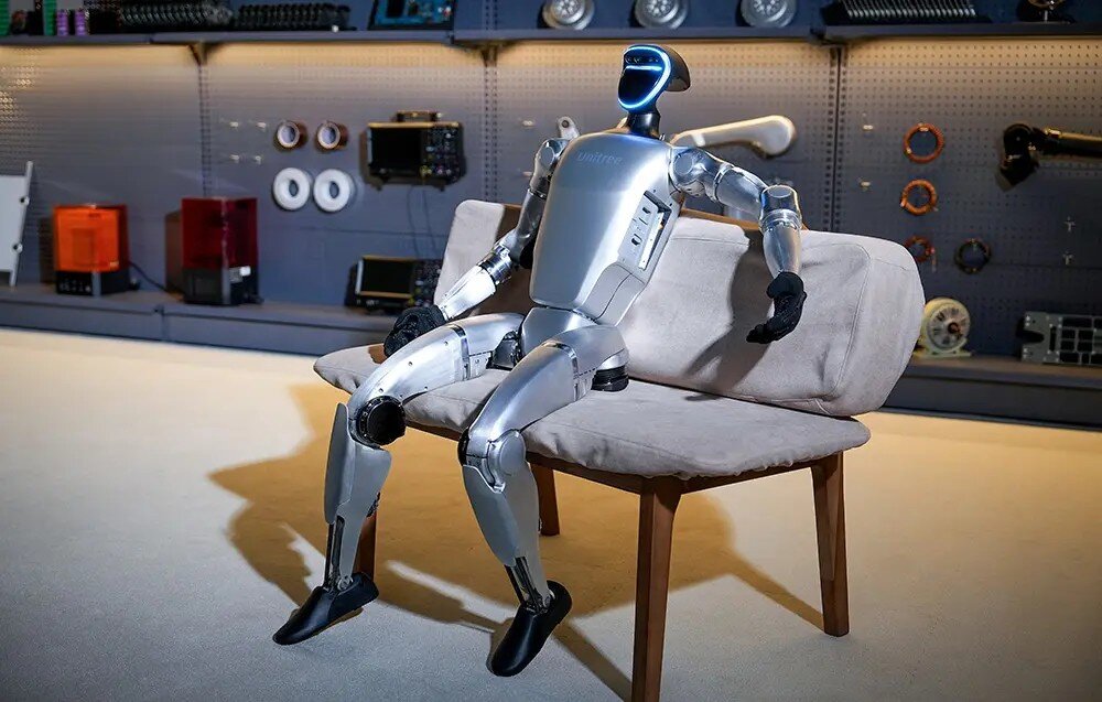 Robot humanoidalny siedzi na sofie w pomieszczeniu z narzędziami i sprzętem elektronicznym na ścianie. Robot ma srebrne elementy i czarny ekran na głowie.