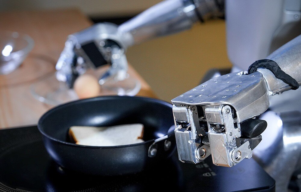 Robot humanoidalny trzyma patelnię z kromką chleba. W tle widoczna jest miska i jajko na stole.