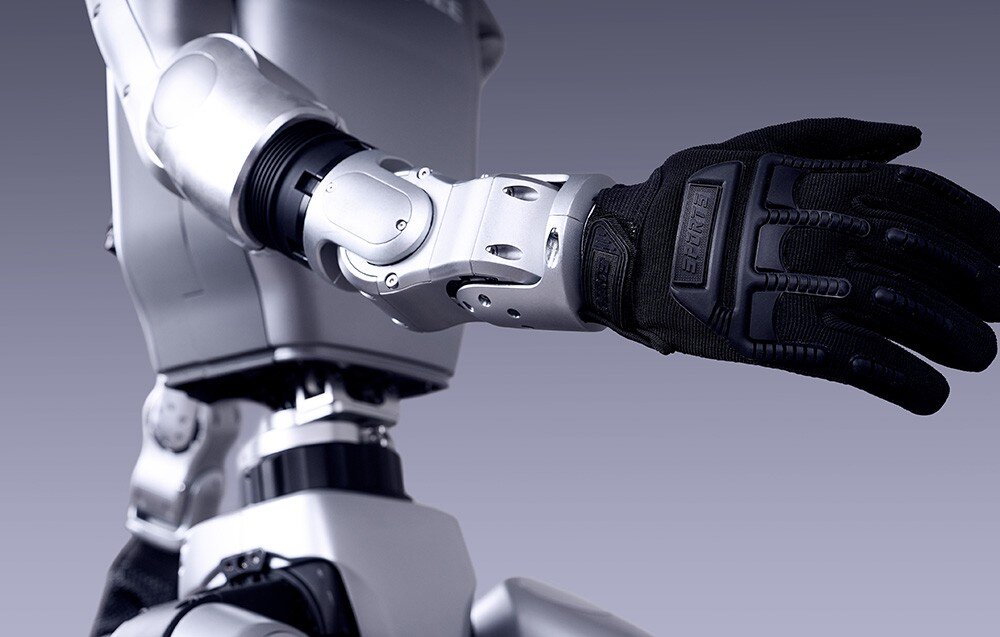 Obraz przedstawia ramię robota z metalicznymi elementami i czarną rękawicą. Tło jest jednolite, w odcieniach szarości.