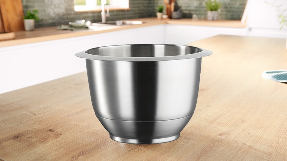 Na drewnianym blacie kuchennym znajduje się metalowa misa o gładkiej powierzchni. W tle widać rozmytą kuchnię z zielonymi roślinami i zlewem.