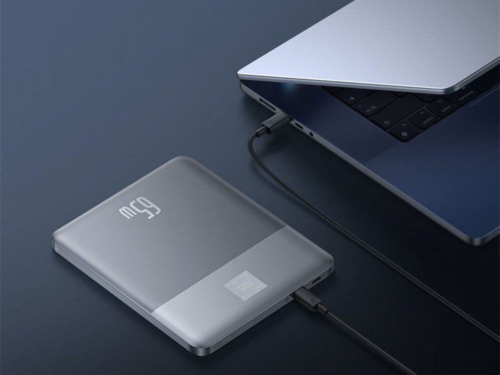 Srebrny powerbank Baseus Blade2 z wyświetlaczem 100% leżący na ciemnym blacie, podłączony czarnym kablem do laptopa
