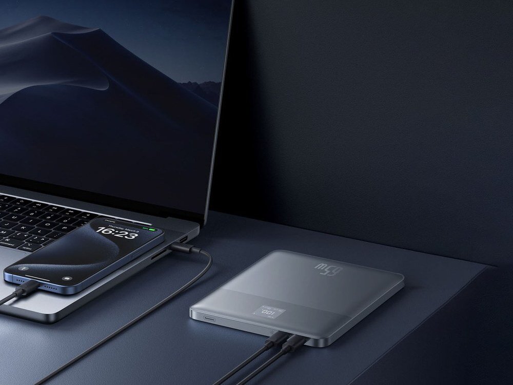 Srebrny powerbank Baseus Blade2 z cyfrowym wyświetlaczem leżący na biurku obok smartfona i laptopa podłączonych kablami.