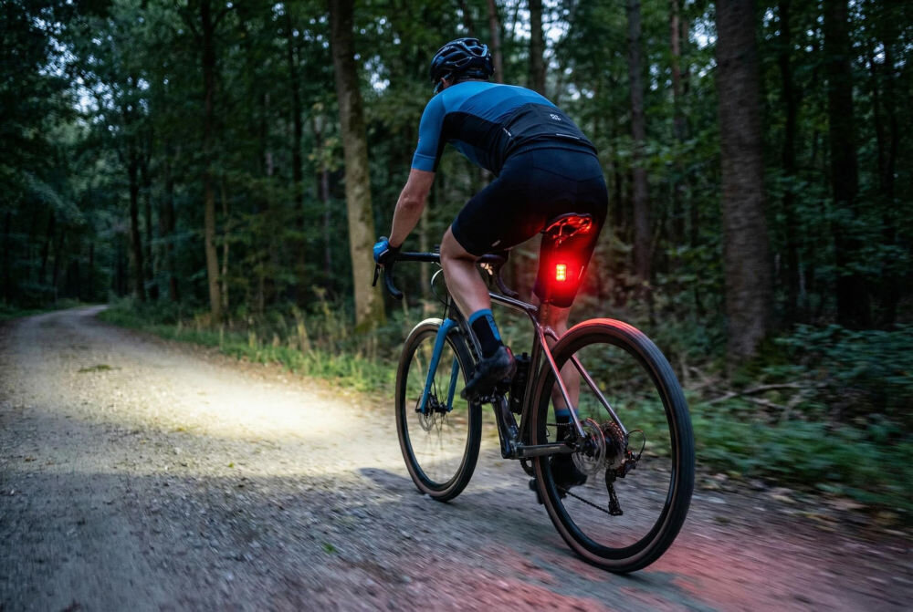 Lampka rowerowa S-LINE BL602 tylna 300 lm czerwone światło akumulator Li-Ion USB-C do jazdy po zmroku Kolarz jadący leśną ścieżką po zmroku z włączonym oświetleniem rowerowym. Zdjęcie ukazuje sylwetkę rowerzysty od tyłu, z wyraźnie widocznym czerwonym światłem tylnym zamontowanym pod siodełkiem oraz mocnym strumieniem białego światła przedniego oświetlającego drogę.