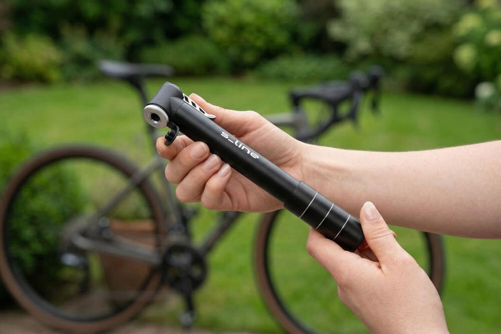 Pompka rowerowa S-LINE Mini VPO120 Kobieta trzyma czarną, teleskopową pompkę rowerową s_line na tle zamazanego roweru szosowego na tle zieleni.