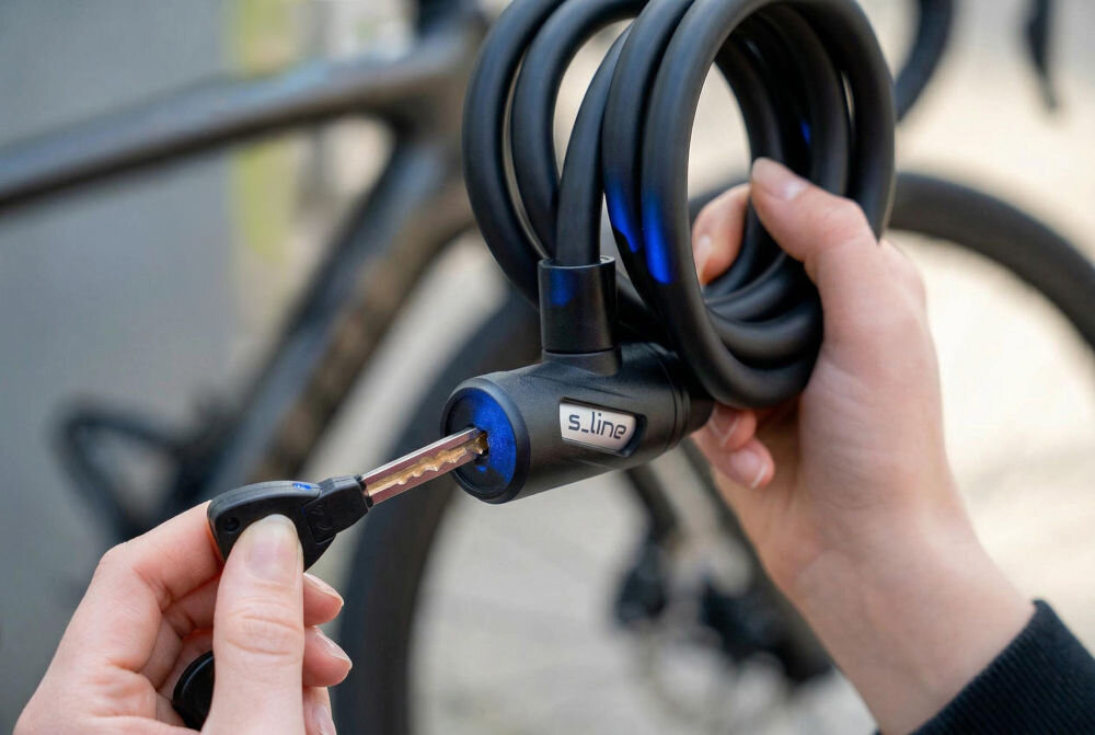 Zapięcie rowerowe S-LINE VZP105 z lampką Linka Czarny Zbliżenie na dłonie wkładające kluczyk do czarnej głowicy zapięcia rowerowego marki s_line, z podświetlonym na niebiesko wnętrzem dziurki na klucz. W tle zamazany rower.