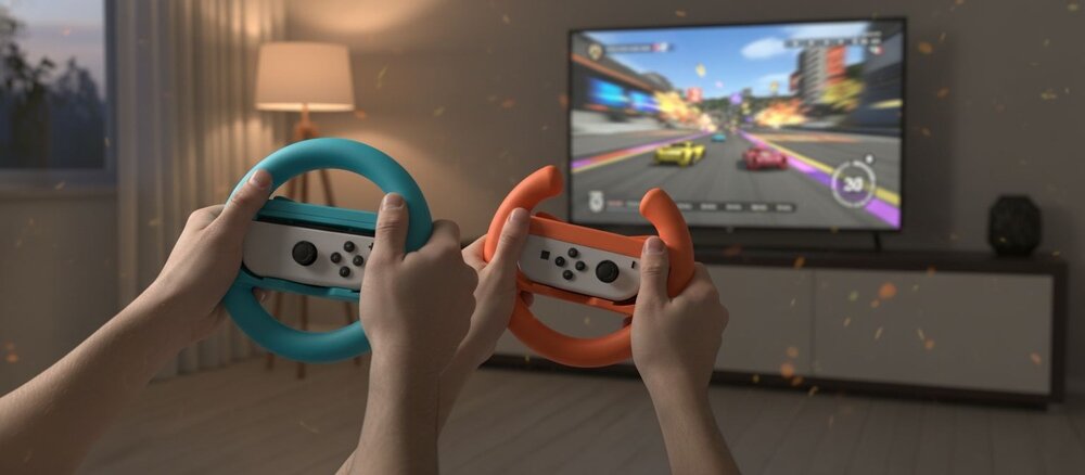Dwie osoby trzymają przed telewizorem niebieską i pomarańczową kierownicę do Joy-Con, na ekranie widać rozmytą grę wyścigową.