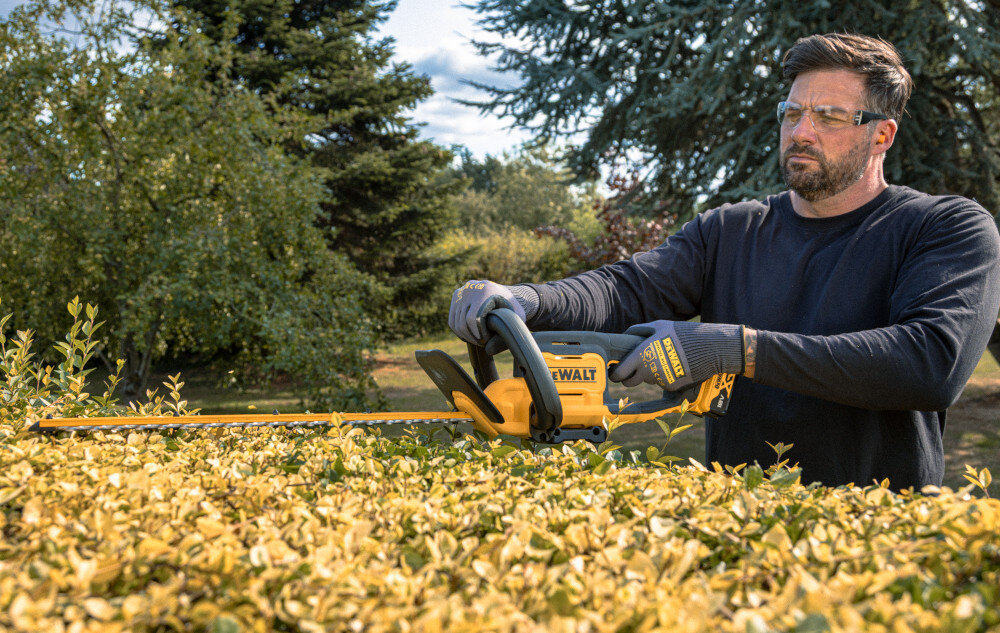 Nożyce do żywopłotu DEWALT DCMHT562P1-QW 550mm 18V akumulatorowe ogród osoba przycina żywopłot w tle trawnik drzewa krzewy niebo pielęgnacja zieleni zadbany ogród sprawne przycinanie żywopłotów krzewów kształtowanie ogrodu