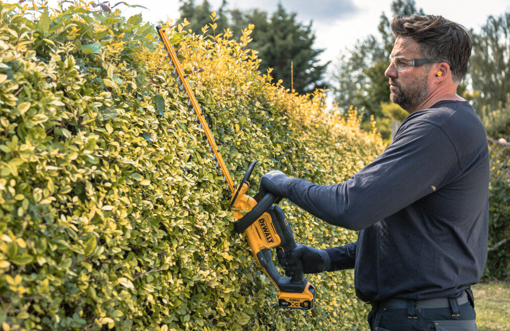Nożyce do żywopłotu DEWALT DCMHT562P1-QW 550mm 18V akumulatorowe ogród osoba przycina żywopłot w tle drzewa niebo do 75 minut na jednym ładowaniu akumulatora ciągłość pracy ma realny wpływ na tempo realizacji wygoda użytkowania