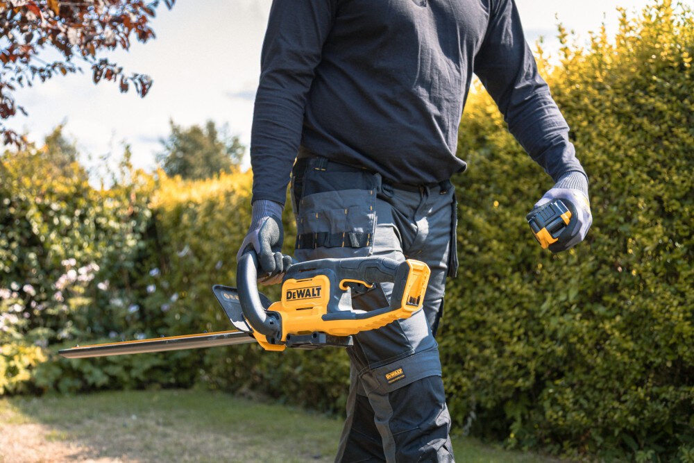 Nożyce do żywopłotu DEWALT DCMHT562P1-QW 550mm 18V akumulatorowe ogród osoba niesie w dłoni nożyce w drugiej dłoni trzyma akumulator w tle żywopłot niebo waga około 3,2 kg bez akumulatora ergonomiczne uchwyty pokryte gumą łatwe prowadzenie narzędzia ogranicza obciążenie ramion pewny chwyt kontrola nad ruchem ostrza