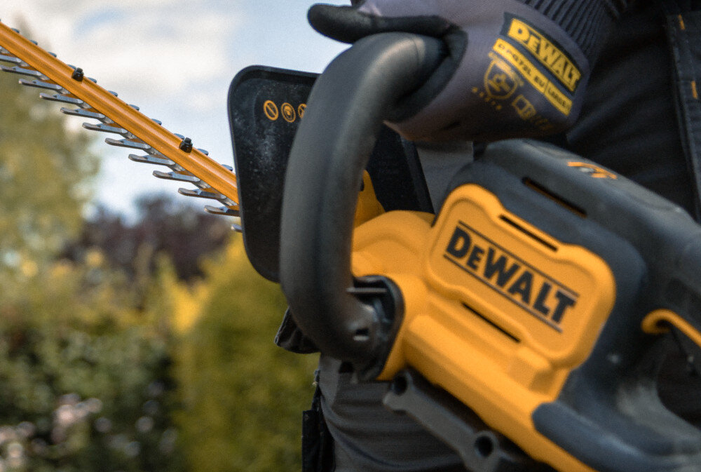 Nożyce do żywopłotu DEWALT DCMHT562P1-QW 550mm 18V akumulatorowe ogród zbliżenie na uchwyt osoba trzyma nożyce w dłoniach rozmazane tło uchwyt w kształcie litery D wygodna obsługa w różnych pozycjach dłoni precyzja pracy ogranicza ryzyko nierównego cięcia swoboda zmiany chwytu