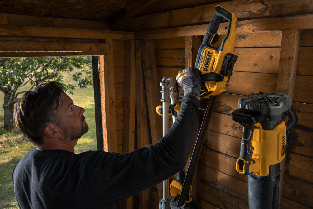 Nożyce do żywopłotu DEWALT DCMHT562P1-QW 550mm 18V akumulatorowe szopa garaż osoba ściąga z drewnianej ściany nożyce obok inne narzędzia ogrodowe hak do montażu na ścianie umieszczony w dolnej części obudowy łatwe przechowywanie porządek lepsza organizacja przestrzeni