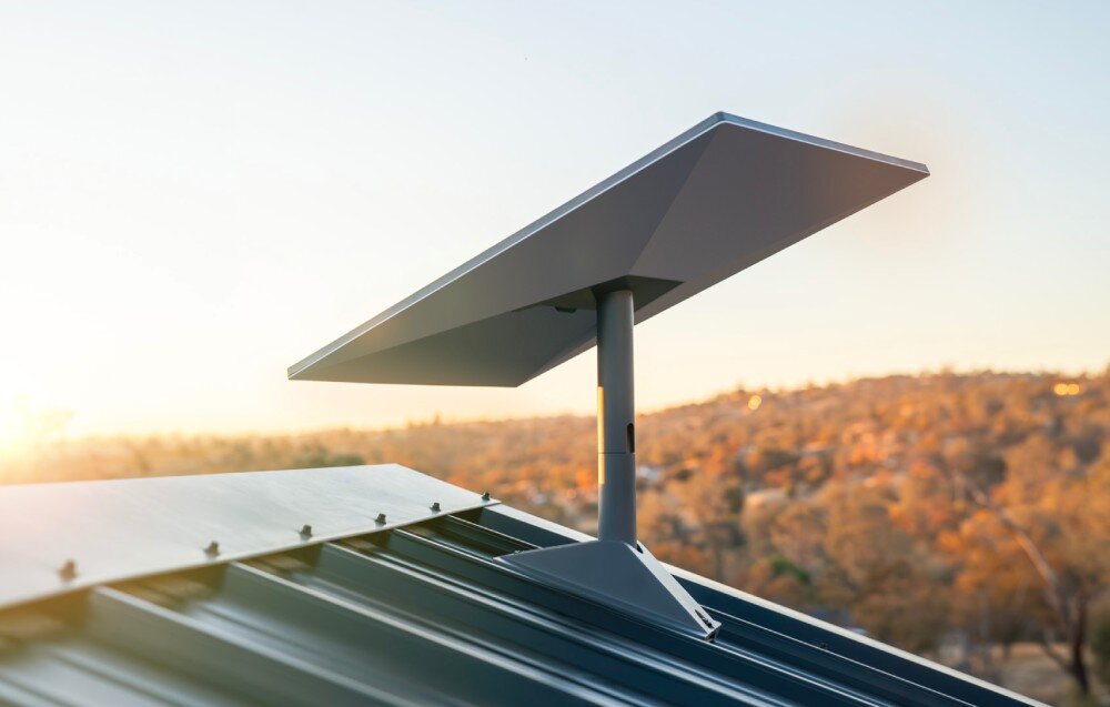 Na dachu budynku zamontowana jest antena satelitarna o nowoczesnym, prostokątnym kształcie. W tle widać krajobraz z drzewami i wzgórzami oświetlonymi przez zachodzące słońce.