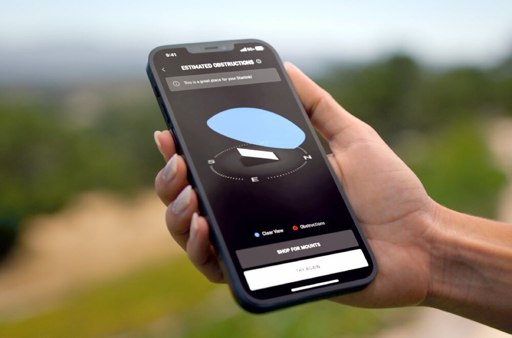 Osoba trzyma smartfon z aplikacją pokazującą mapę przeszkód dla Starlink. Na ekranie widoczny jest tekst: \'ESTIMATED OBSTRUCTIONS\' oraz przyciski \'SHOP FOR MOUNTS\' i \'TEST AGAIN\'.
