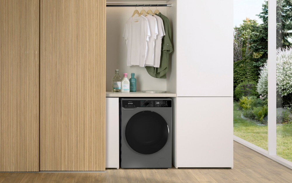 Pralko-suszarka GORENJE G600 WD2PA964ADT/PL W niszy zabudowy umieszczono ciemnoszarą pralko–suszarkę ładowaną od frontu, nad którą znajduje się blat z butelkami detergentów. Powyżej wisi drążek z kilkoma koszulami na wieszakach, a po prawej stronie przez wysokie okno widać zielony ogród, co nadaje aranżacji jasny, naturalny charakter