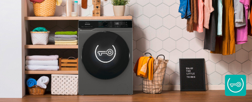 Pralko-suszarka GORENJE G600 WD2PA964ADT/PL W przytulnej pralni ustawiono ciemnoszarą pralko–suszarkę z dużymi, czarnymi drzwiami, na których widnieje podświetlona ikona klucza symbolizująca blokadę bezpieczeństwa. Po prawej stronie stoi kosz z ubraniami i tablica z napisem „ENJOY THE LITTLE THINGS”, a w prawym dolnym rogu znajduje się turkusowa ikonka klucza, podkreślająca funkcję blokady przed dziećmi ChildLock blokada panelu bezpieczeństwo dzieci ochrona ustawienia aktywacja dezaktywacja