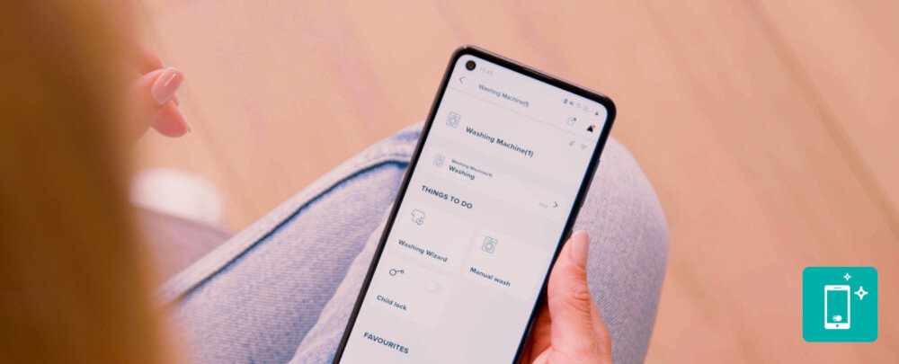 Pralko-suszarka GORENJE G600 WD2PA964ADT/PL Kobieta siedząca po turecku trzyma w dłoni smartfon z aplikacją do obsługi pralki, w której widać status urządzenia i dostępne funkcje, takie jak zdalne uruchamianie czy blokada rodzicielska. W prawym dolnym rogu umieszczono turkusową ikonkę telefonu ze świecącymi gwiazdkami, podkreślającą wygodne, mobilne sterowanie sprzętem ConnectLife WiFi sterowanie pralka kreator programów tkaniny plamy ustawienia komfort skuteczność
