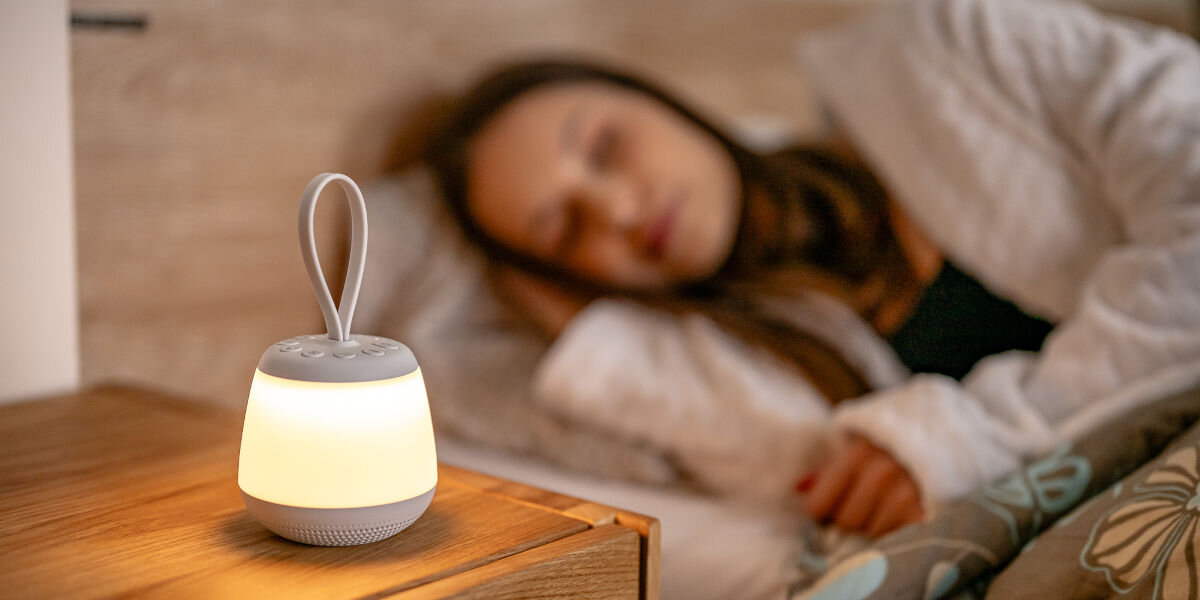Lampka nocna LED NEWELL SeleneDream NL4496, biały, szum, sen, wyciszenie, Zapalona lampka nocna LED Newell SeleneDream stoi przy łóżku na drewnianej szafce nocnej. W tle śpiąca kobieta.