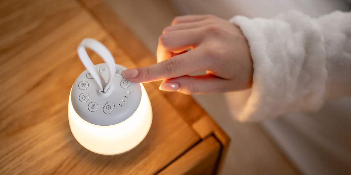 Lampka nocna LED NEWELL SeleneDream NL4496, obsługa, przyciski, intuicja, wygoda, Kobiecy palec wskazujący naciska wyłącznik zapalonej lampki Newell SeleneDream. Lampka stoi na drewnianej szafce nocnej.