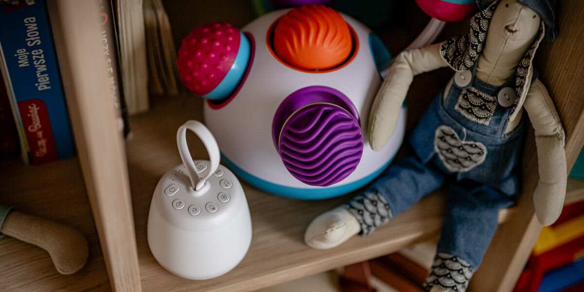 Lampka nocna LED NEWELL SeleneDream NL4496, design, stabilność, funkcjonalność, bezpieczeństwo, Lampka nocna LED Newell SeleneDream z funkcją białego szumu na drewnianym regale pośród innych zabawek i książek.