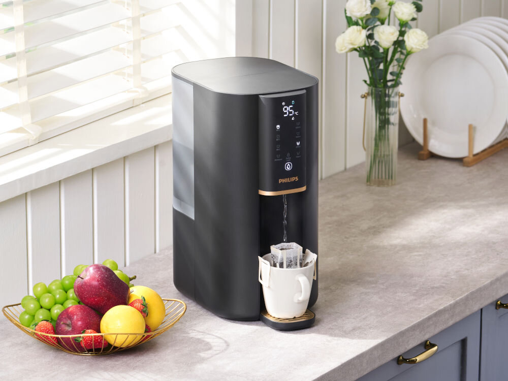 Dyspenser do wody PHILIPS ADD6901HBK01/10 Nowoczesny dystrybutor wody Philips z wyświetlaczem temperatury na blacie kuchennym, obok kosza z owocami i wazonu z białymi kwiatami.