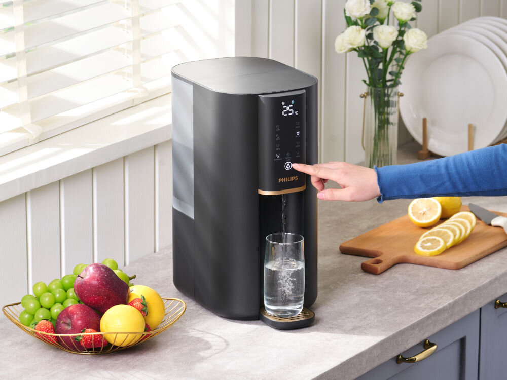 Dyspenser do wody PHILIPS ADD6901HBK01/10 Dłoń naciskająca przycisk na panelu dotykowym dystrybutora wody Philips, obok szklanki wody i deski do krojenia z cytrynami.