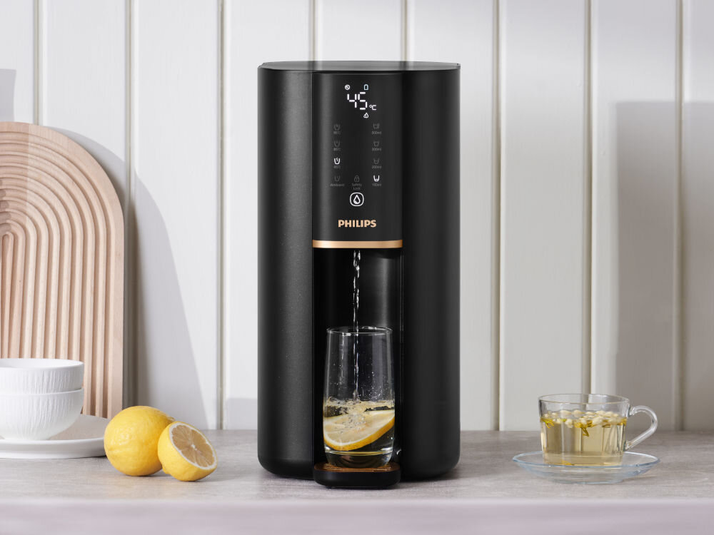 Dyspenser do wody PHILIPS ADD6901HBK01/10 Dystrybutor wody Philips z przodu, z podświetlonym wyświetlaczem cyfrowym na tle białych paneli ściennych, z cytrynami i filiżanką herbaty.