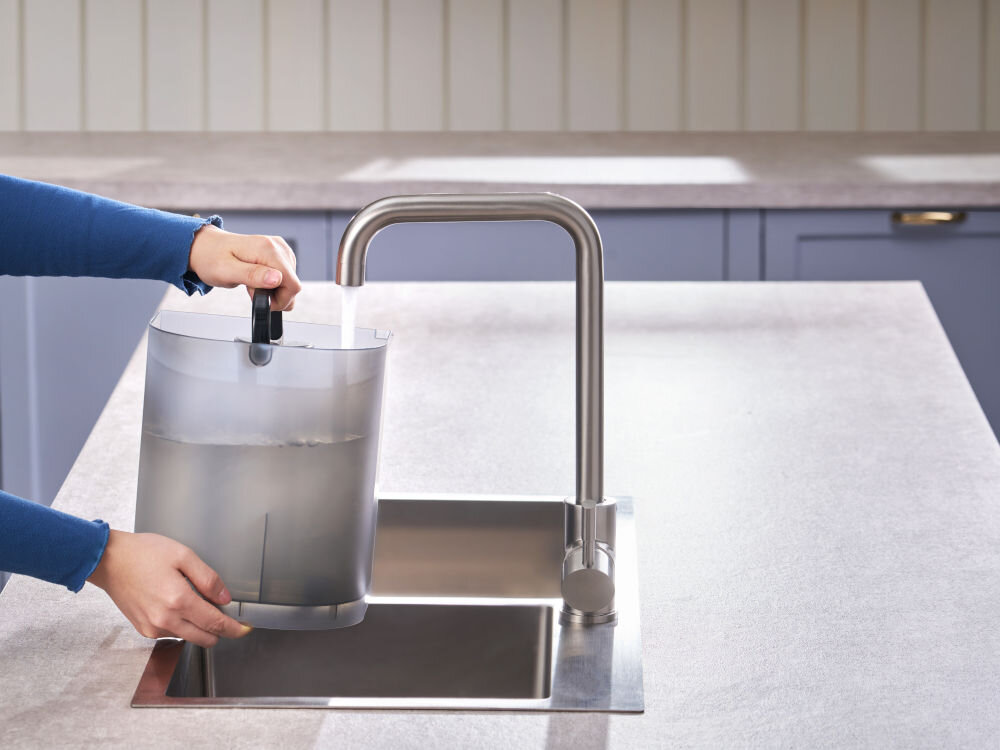 Dyspenser do wody PHILIPS ADD6901HBK01/10 Dłonie trzymające zbiornik na wodę pod strumieniem wody z kranu kuchennego nad zlewozmywakiem.